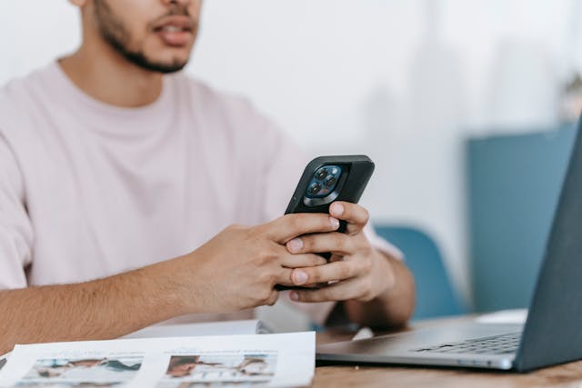 Čovek drži mobilni telefon u rukama na stolu pored laptopa